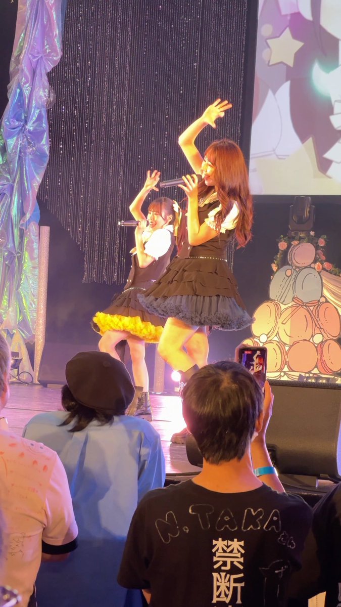 Two female performers in black dresses singing on stage, with audience members visible in the foreground, including one wearing a 'N.TAKA' shirt with Japanese characters.