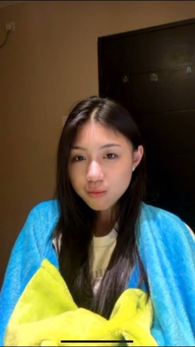 A young woman with long dark hair poses for a selfie, wrapped snugly in a bright blue and yellow blanket while wearing a white shirt. The cozy, bundled-up scene perfectly captures her playful post about being cold from the AC yet too lazy to turn it off, with the colorful blanket serving as her makeshift defense against the chill.
