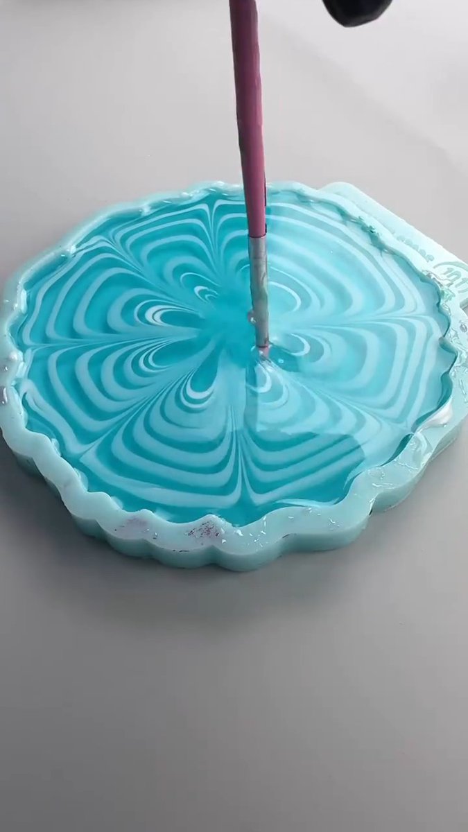 A close-up shot captures a stick stirring swirling blue and white paint inside a small, flower-shaped dish, creating a marbled pattern.