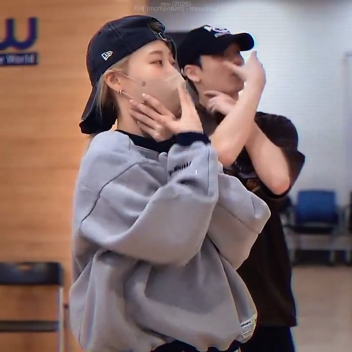 The image shows two people wearing caps and masks, appearing to be in a dance practice studio, likely during a rehearsal for Moonbyul's 2026 song "MOMENTUM" as indicated by the text "momentum - moonbyul" visible in the upper right corner. The person in the foreground wears a gray oversized sweatshirt and a backward cap, with their hand raised near their face in a pose that suggests dance practice or choreography preparation.