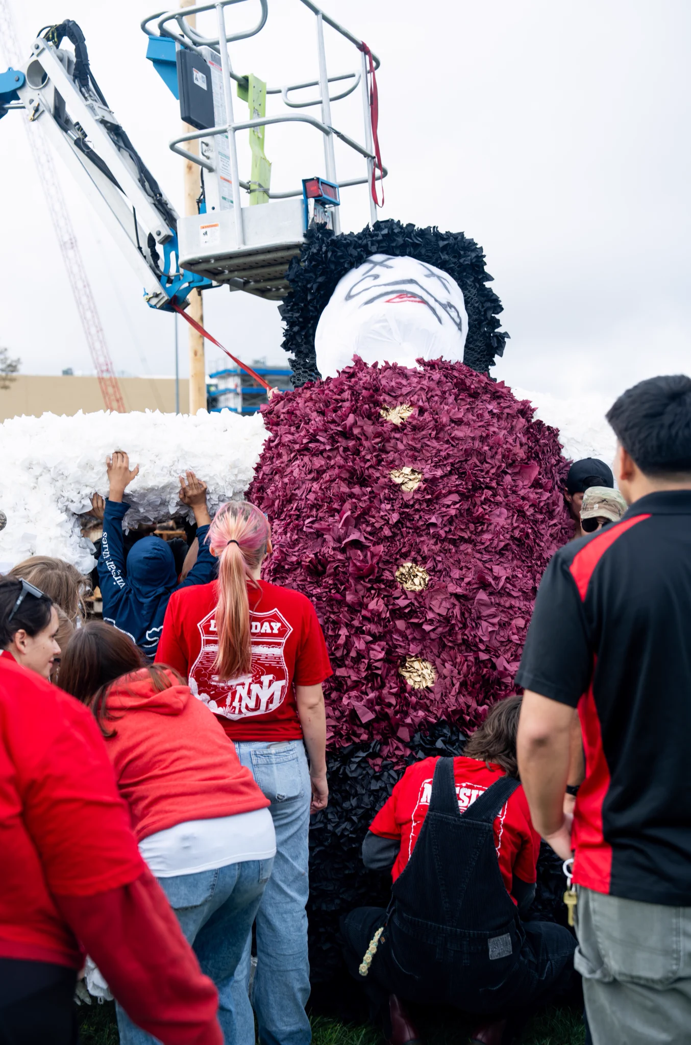 A large Aggie effigy is being held up by a group of people wearing UNM merch. A crane is visible in the background.