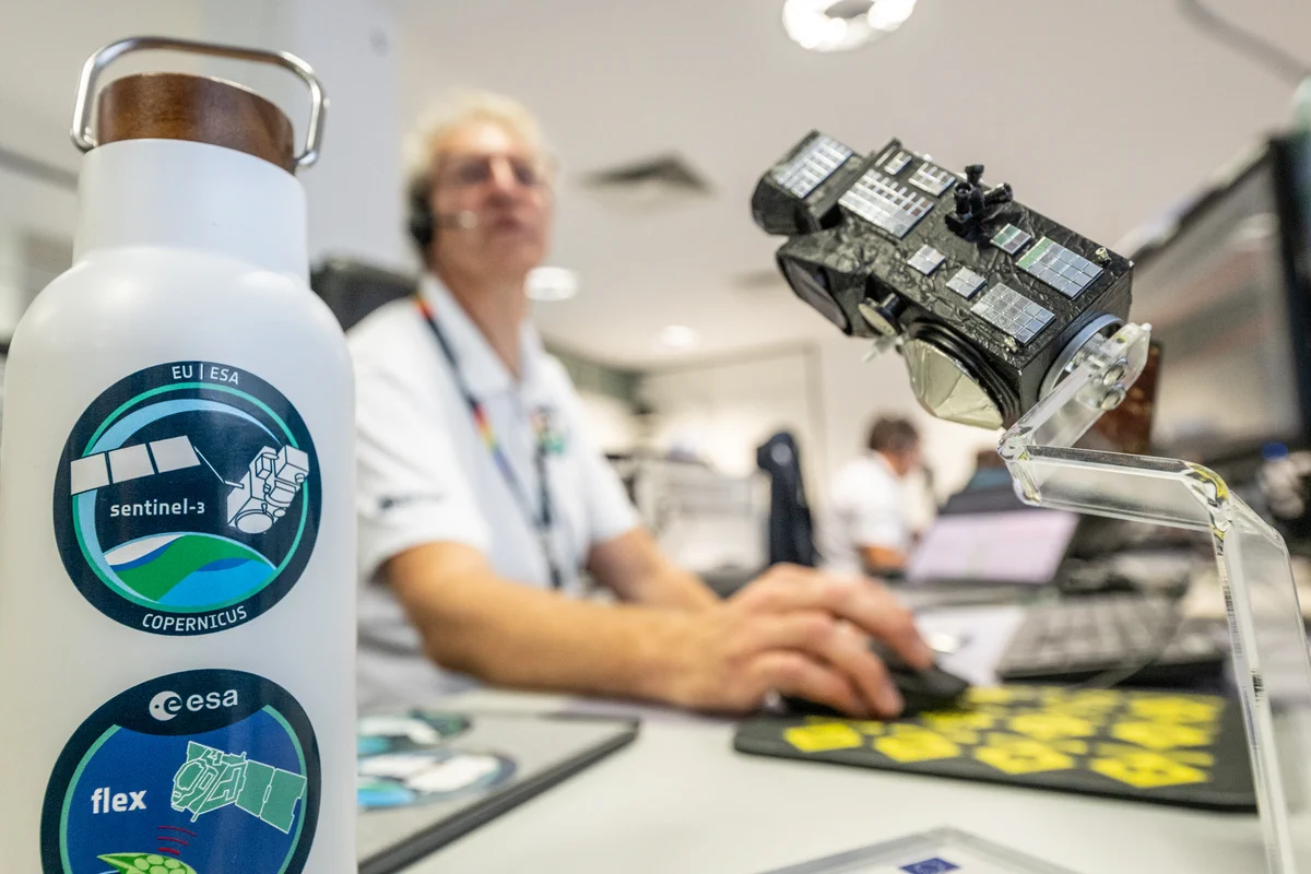 Close-up of a Sentinel‑3 mission sticker and FLEX mission sticker on a water bottle beside a small satellite model on a desk in a mission operations room at ESA’s European Space Operations Centre (ESOC) during the launch and early orbit phase (LEOP) for Copernicus Sentinel-3C.

In the background, Franco Marchese, ESA Flight Operations Director for the Sentinel-3C LEOP, is wearing a headset works at a computer workstation, with screens and control-room equipment visible out of focus.