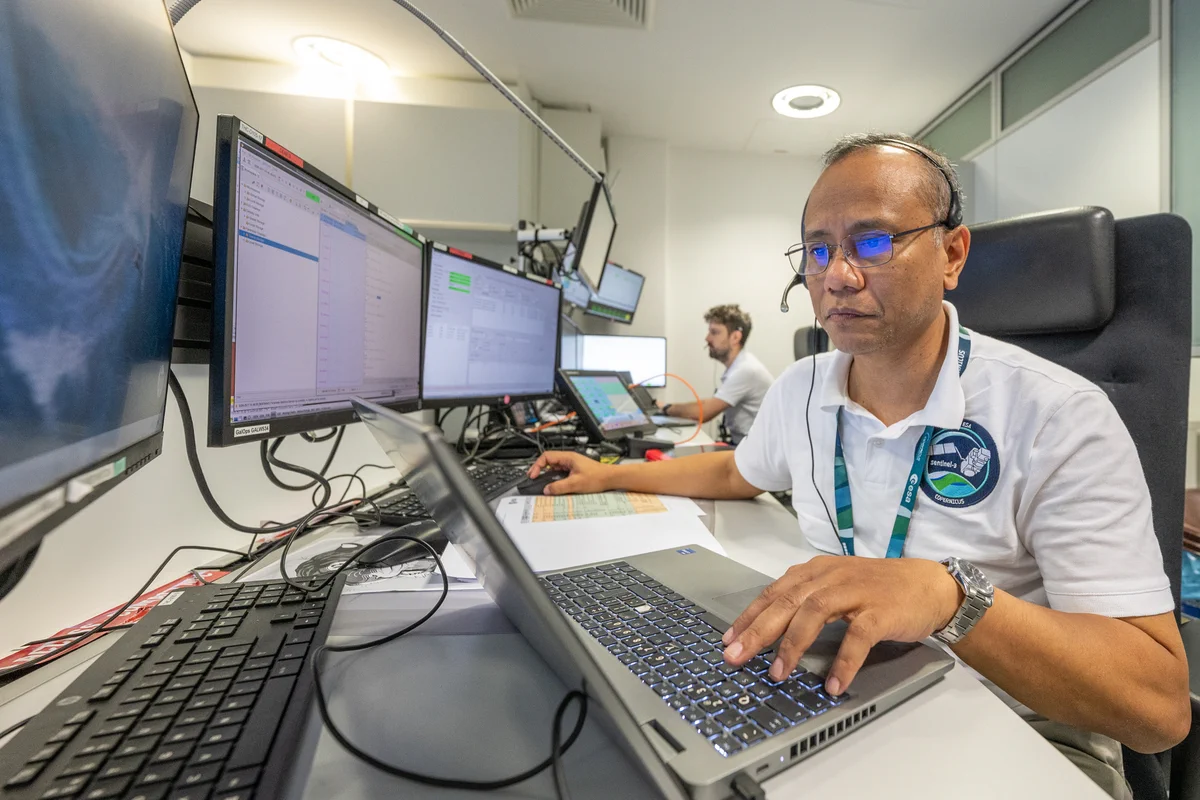 Two Ground Operations Managers, Pablo Asenjo Garcia and Ruddy-Herard Chatim, sit at a workstation at ESA’s European Space Operations Centre (ESOC) during the launch and early orbit phase (LEOP) for Copernicus Sentinel-3C.

The workstation features multiple monitors displaying spacecraft operations data and communication schedules. Laptops, consoles, and control-room equipment are arranged across the desk, while additional screens and workstations fill the background of the mission operations room.