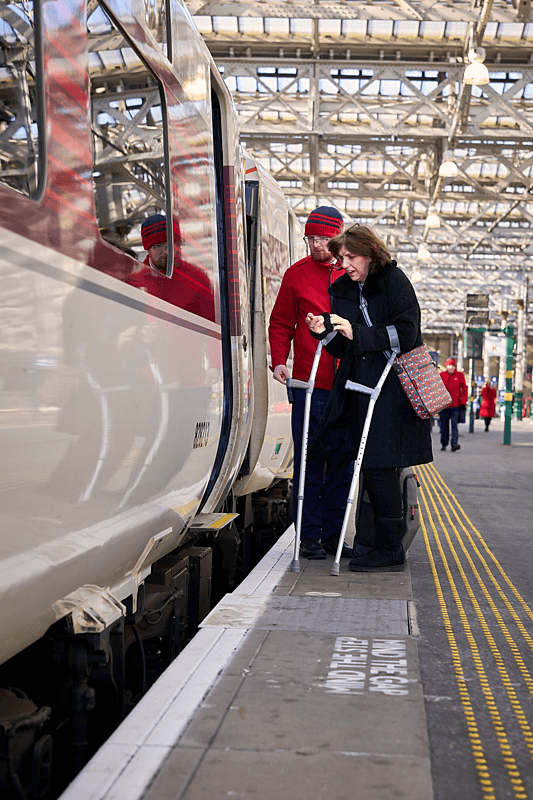 A person with crutches is assisted onto a train by  a LNER member of staff  at a train station. The platform has yellow safety lines, and the station features a large arched glass roof.