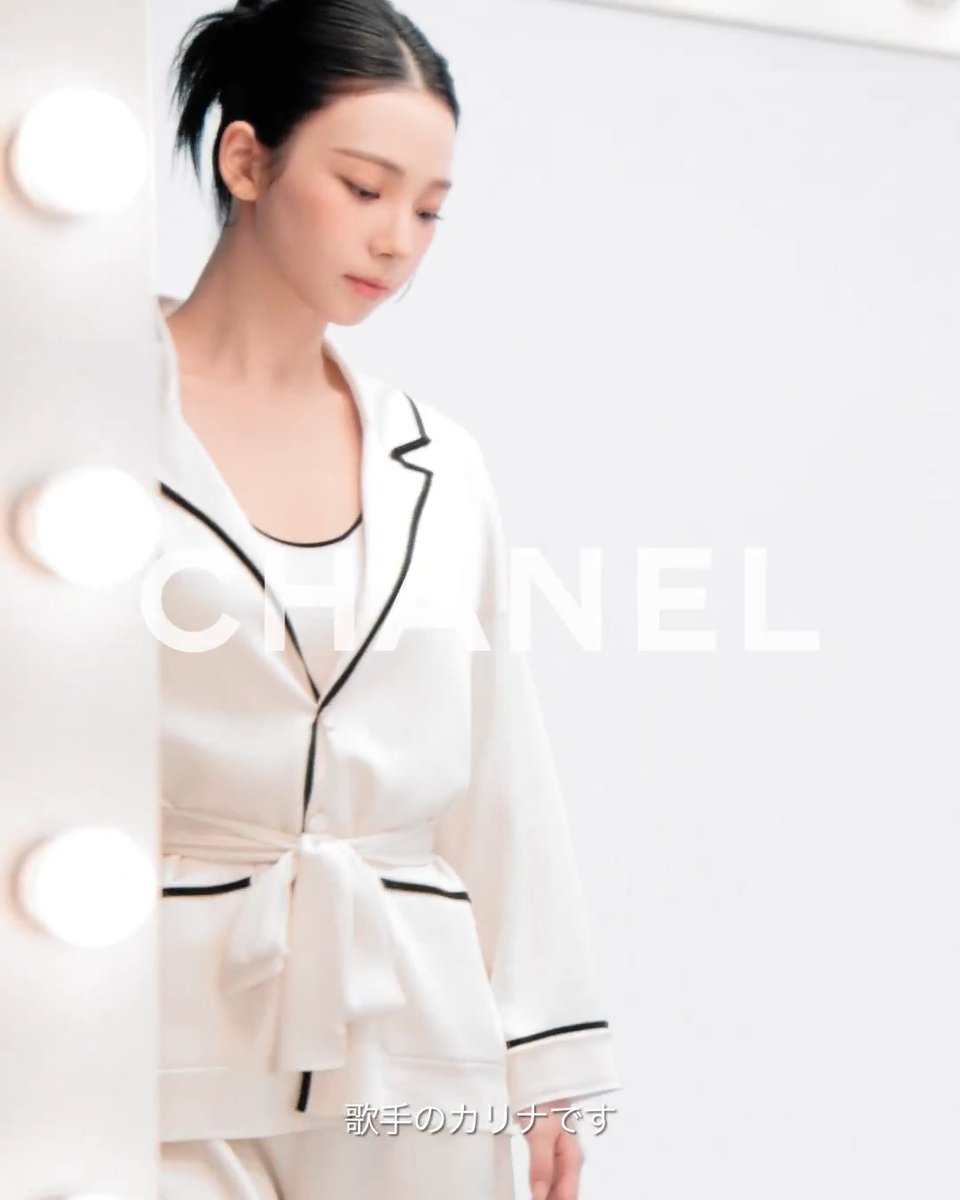 A Japanese woman wearing white Chanel pajamas with black piping stands in a brightly lit dressing room with Hollywood-style mirror bulbs, with Japanese text reading '歌手のカリナです' (I am Karina, a singer).