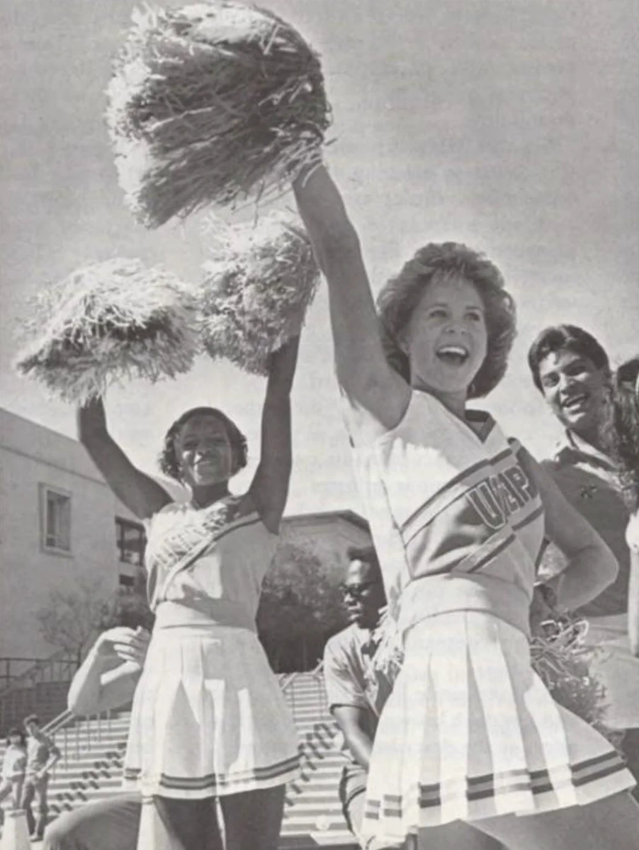 The image shows cheerleaders from UTEP performing with pom-poms raised. They are outdoors with a building steps in the background.
