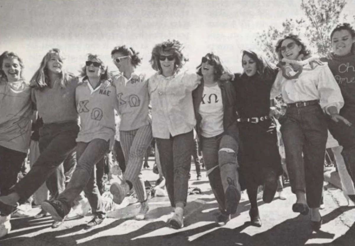 The Chi Omega sisters, standing outdoors, with arms around each other, are joyfully lifting their legs in a coordinated manner at the 1986 UTEP Homecoming pep rally. Some wear shirts with the Greek letters "ΧΩ" visible. Trees can be seen in the background.
