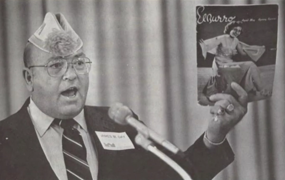 A person speaks into a microphone while holding up a copy of "El Burro" magazine, which features a cover image of a performer. The individual wears a suit, tie, and a hat made from a paper bag. A name tag is visible on the suit.
