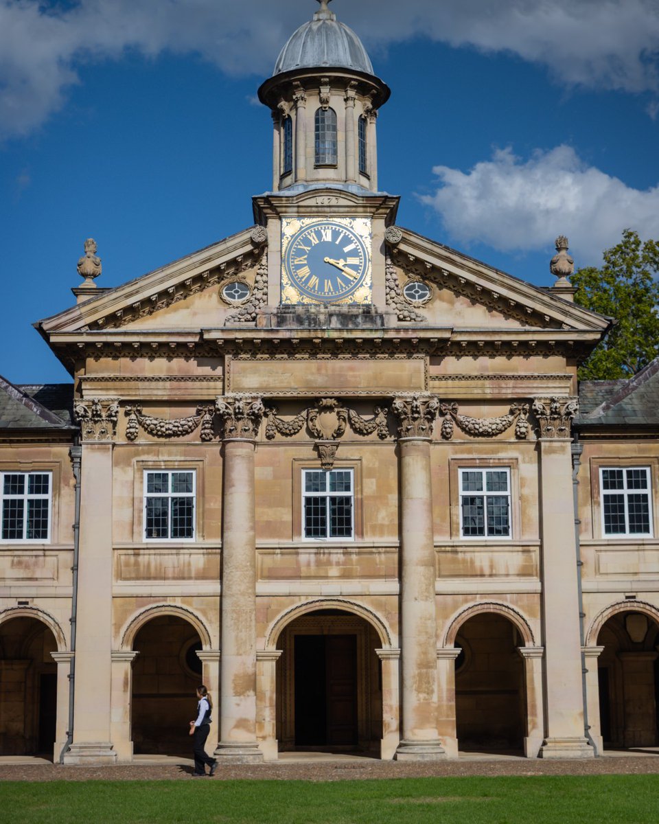 A person walks in front of an ornate, historic building with a clock tower at Emmanuel College, Cambridge. The façade features columns and arches, set against a backdrop of blue sky and clouds.