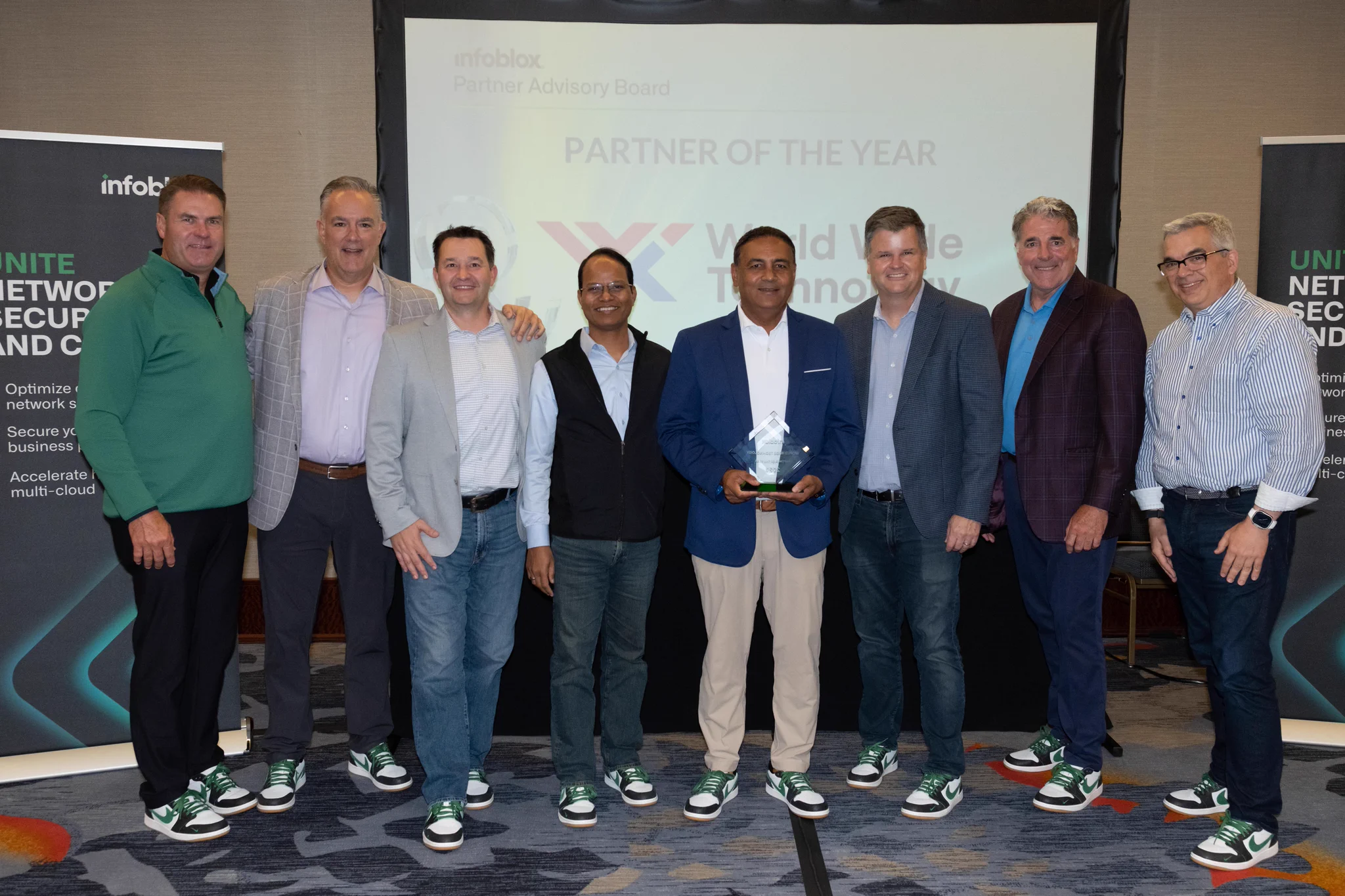 Eight smiling men, including one holding a clear award, pose for a group photo. All wear identical black, white, and green sneakers. A screen behind them reads \"infoblox Partner Advisory Board PARTNER OF THE YEAR World Wide Technology.\"