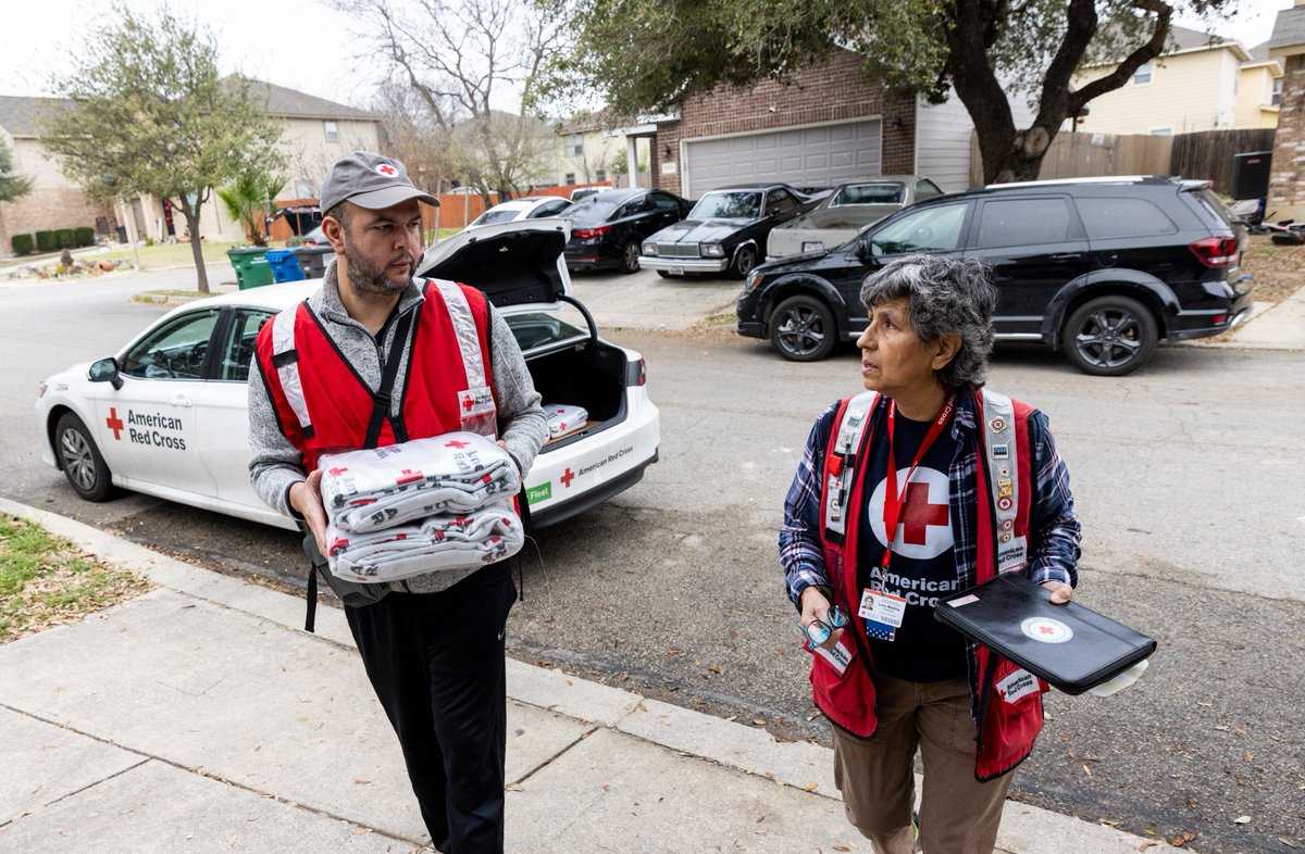 Two people wearing American Red Cross vests are walking on a residential street. One is carrying blankets and the other is holding a clipboard. An American Red Cross vehicle is parked nearby.