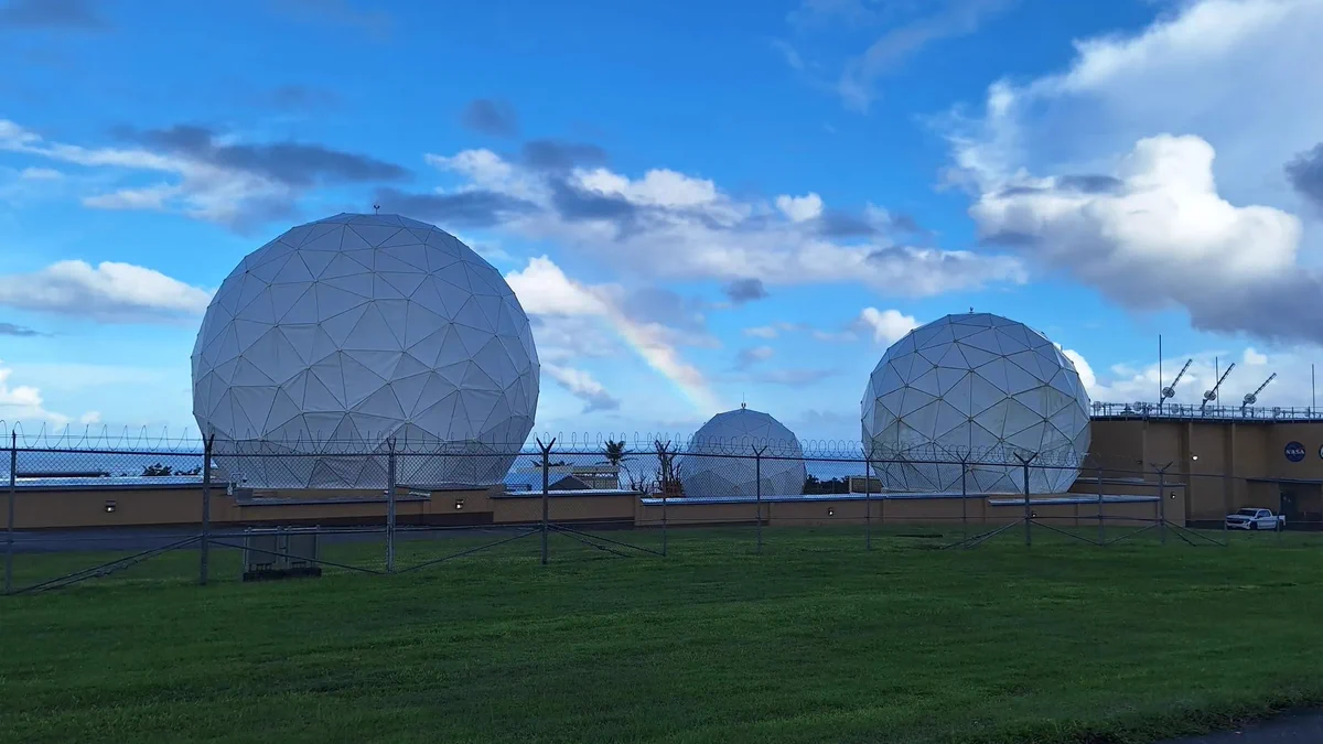 Three large white spherical radio antennas at the repaired Guam Remote Station stand behind a security fence on a grassy field. A rainbow arcs through the partly cloudy blue sky behind the antennas.
