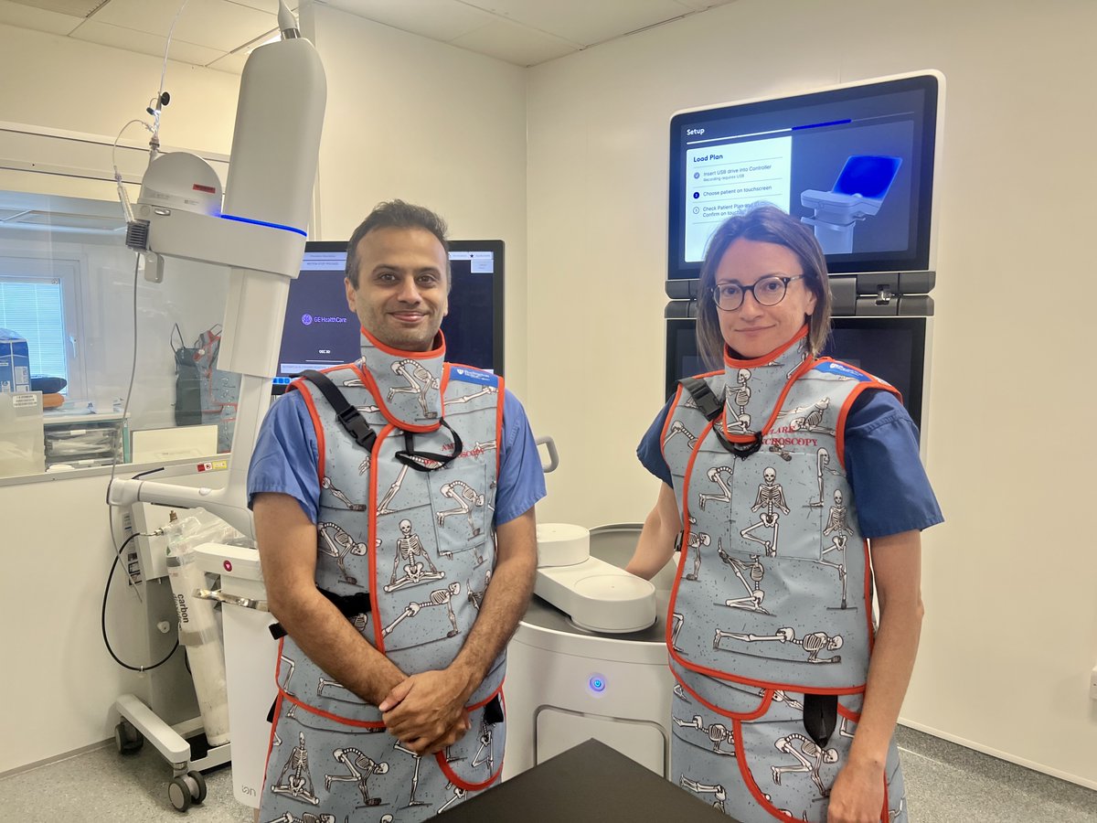 Two medical professionals wearing protective lead aprons with skeleton illustrations stand in a radiology room with imaging equipment behind them.
