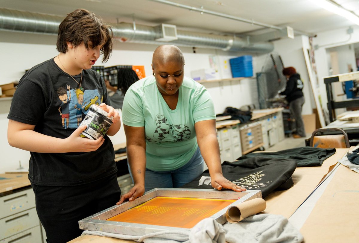 A person assists another in a printmaking studio, showing how to use a screen printing frame. Various materials, including a bottle of ink and clothing items, are scattered on the table.