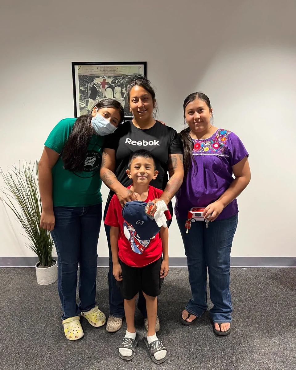 A group of four people standing indoors in front of a framed picture. One person is wearing a Reebok shirt, and another is wearing a mask. A plant is visible to the side.