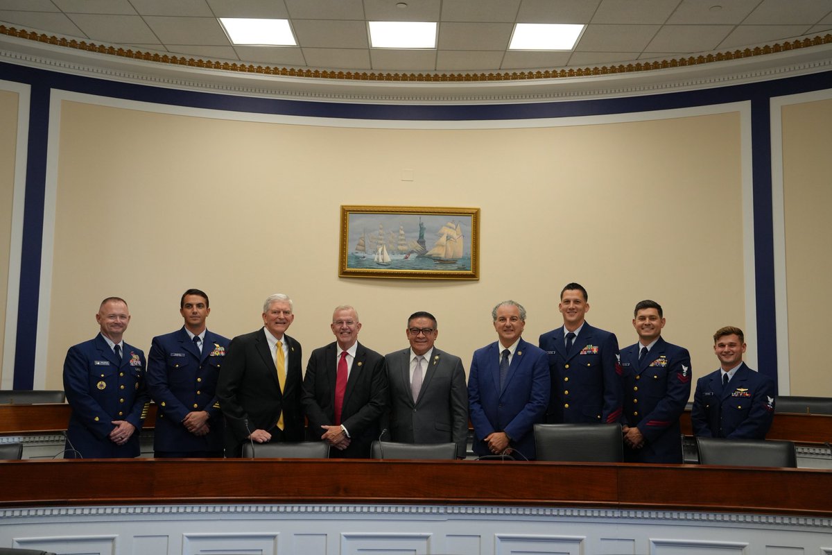 A group of nine men, including several in military uniforms and several in business suits, pose for a photograph behind a curved wooden conference desk in a formal room. A framed painting of sailing ships hangs on the wall behind them. Some clothing and uniform insignia contain text.