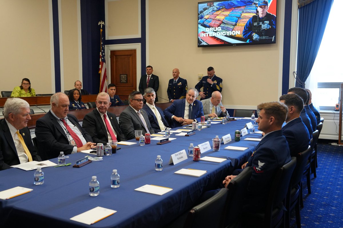 A group of officials and uniformed personnel sit around a long conference table in a formal meeting room. A television displays an image related to drug interdiction. There is text on the television and on nameplates.