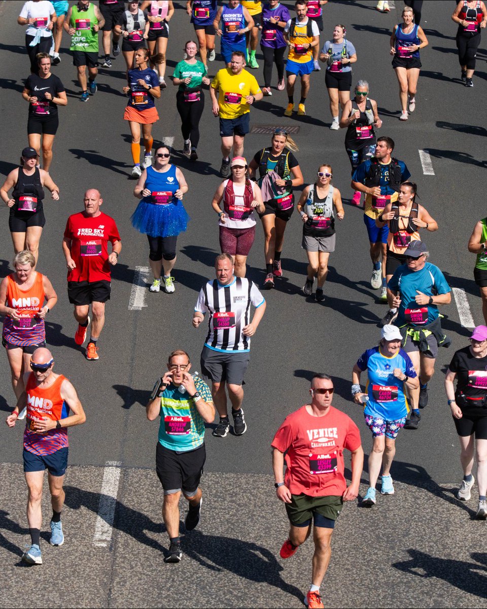 A diverse group of marathon runners in colorful attire is participating in the 2023 TCS London Marathon, moving along a city road. Participants wear various running gear, and some display charity and sponsor logos on their shirts. Spectators are visible in the background.