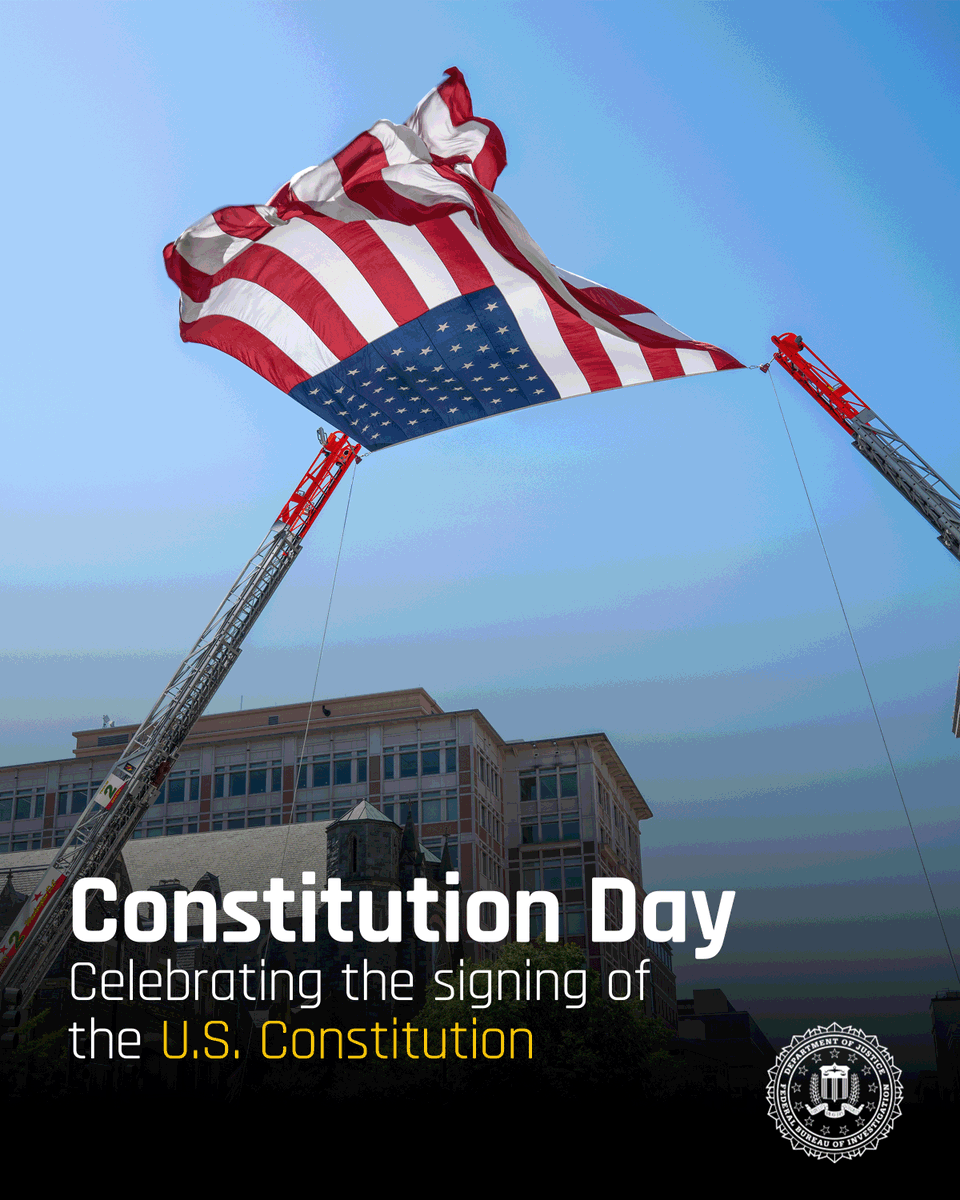 Large American flag hanging between modern buildings under a clear blue sky, symbolizing Constitution Day.