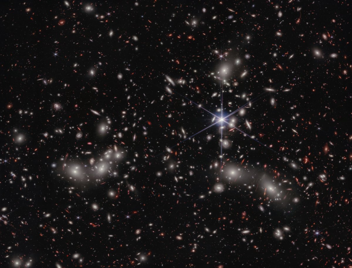 A crowded galaxy field on a black background, with one large star dominating just right of center. Three areas are concentrated with larger white hazy blobs on the left, lower right, and upper right above the single star. Scattered between these areas are many smaller sources of light; some also have a hazy white glow, while many others are red or orange. Even without zooming in, different galaxy shapes are detectable, like spirals, ovals, and arcs.