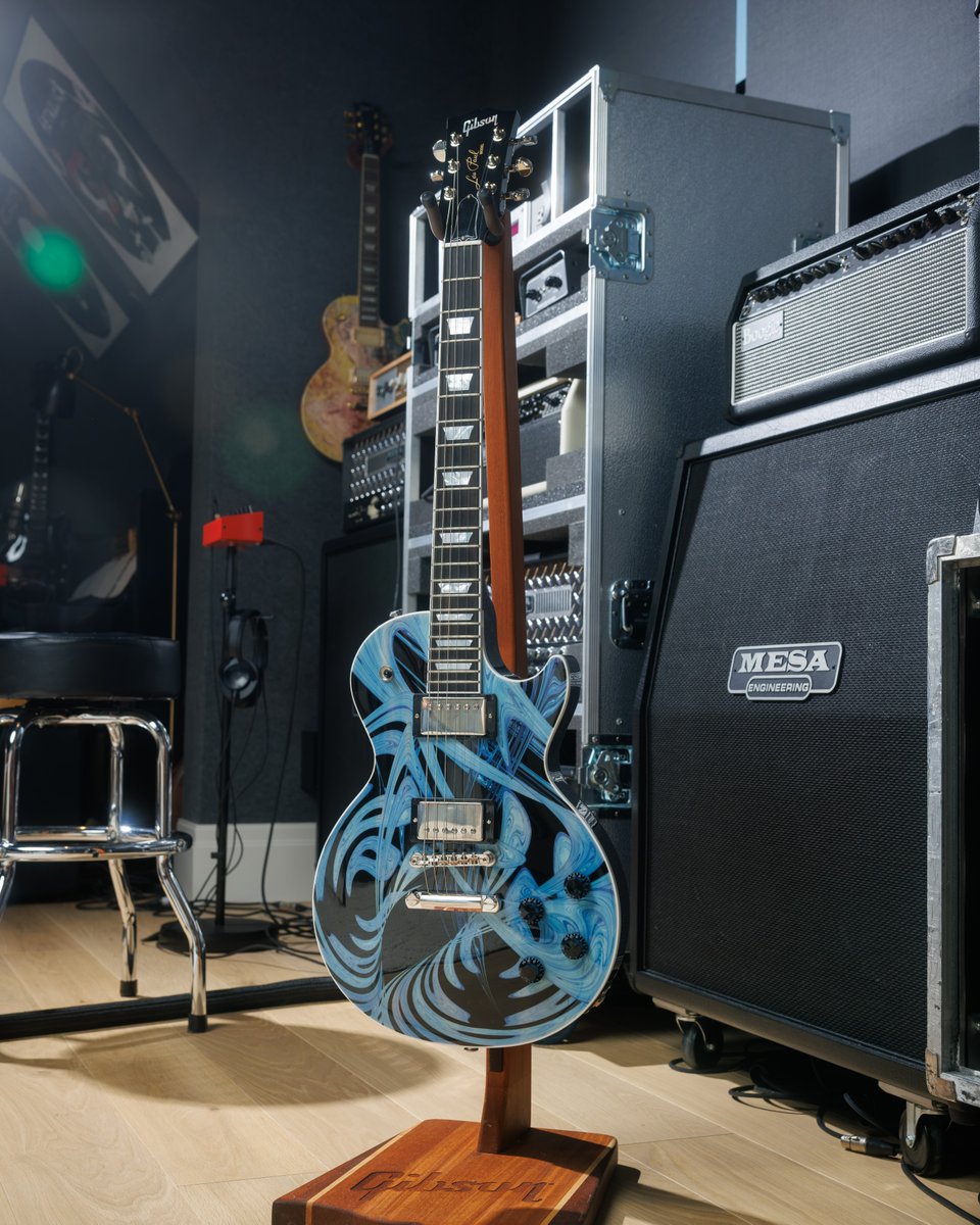 A blue and black one-of-one Gibson Les Paul electric guitar with intricate swirl designs that is artwork created from the soundwaves from the Gibson Band single "Sanctuary" is displayed on a stand next to a Mesa Engineering amplifier in a music studio. The room features additional musical equipment and a stool. A framed guitar is visible on the wall.