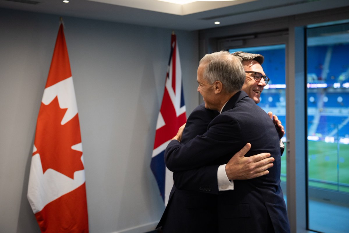 Prime Minister Mark Carney and the Prime Minister of the United Kingdom, Andy Burnham, hug during a meeting at Hill Dickinson Stadium in Liverpool, United Kingdom. The flags of Canada and the United Kingdom are behind them.