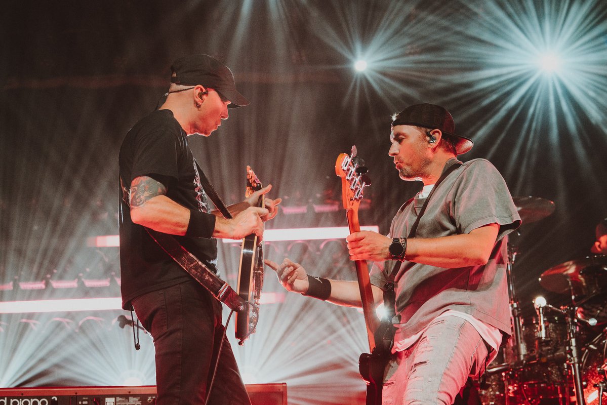 Two musicians playing guitars with bright stage lights behind them during a concert.