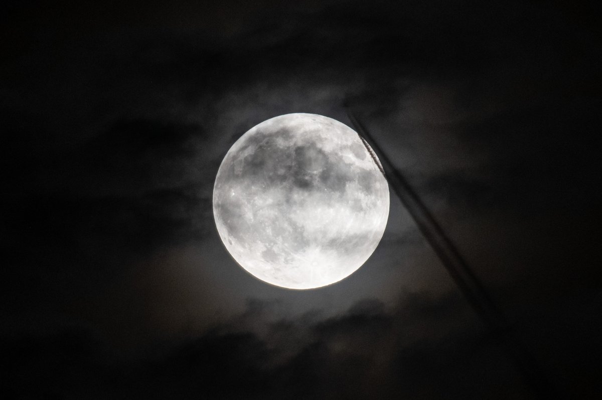 A black-and-white view of a full Moon, partially obscured by clouds and an aircraft trail. Credit: NASA/Jordan Cochran
