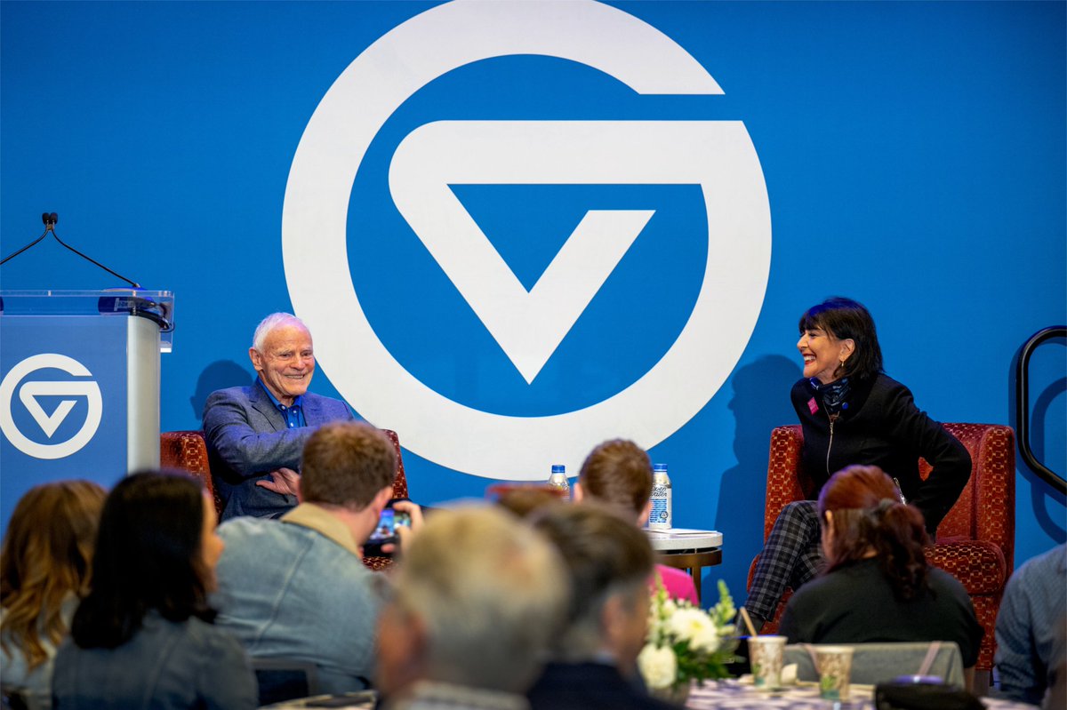 Leroy Hood and President Mantella sit on a stage in front of a large GVSU logo backdrop and have a conversation.