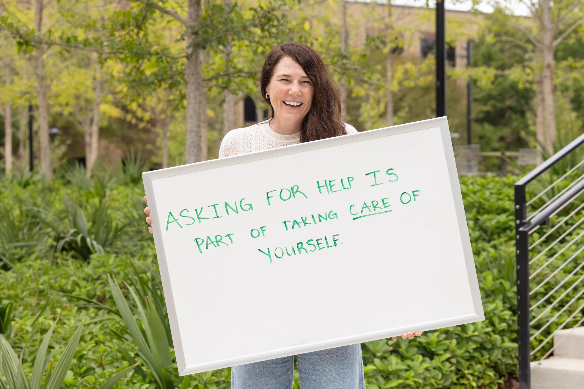 A professor holds up a whiteboard at Wilhelmina's Grove that reads "Asking for help is part of taking care of yourself."