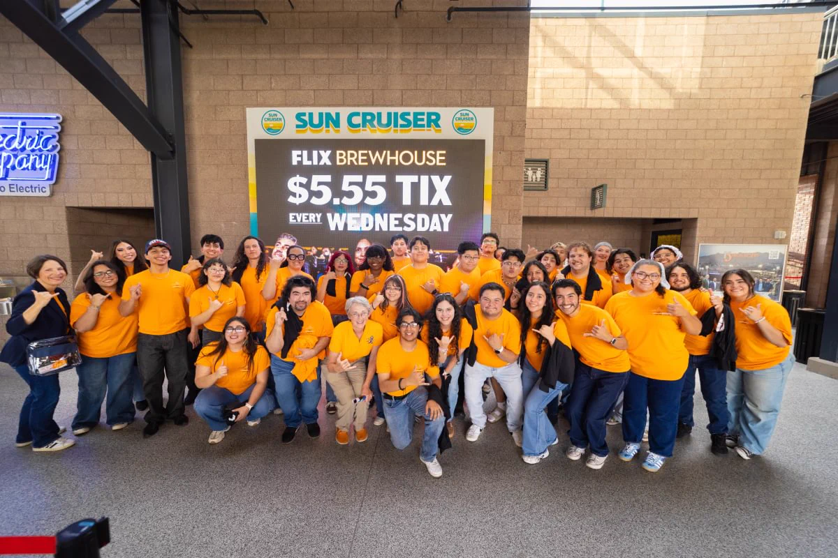 A large group of people wearing matching orange UTEP shirts pose together in an outdoor area in Southwest University Park. 