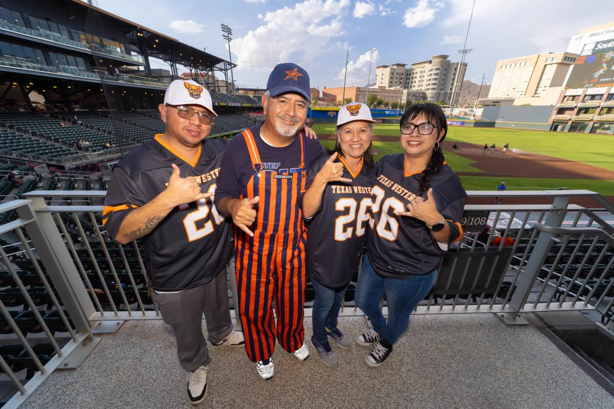 Four individuals are standing together at a baseball stadium. Three are wearing Texas Western jerseys and one is in striped orange. The field and seating areas are visible in the background.
