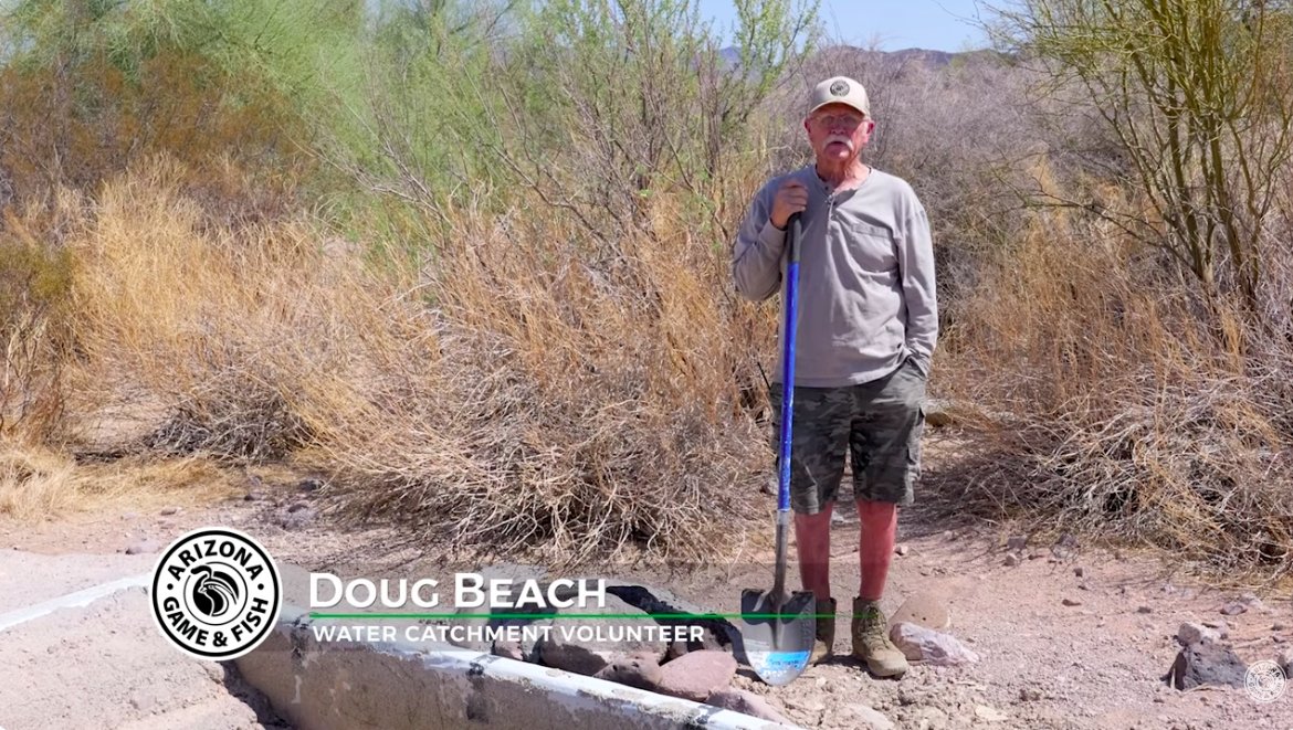 An individual standing outdoors beside a water catchment area, holding a shovel. They're wearing a cap and casual clothing in a desert-like environment. The text on the image reads "Doug Beach, Water Catchment Volunteer." The Arizona Game & Fish logo is also visible.