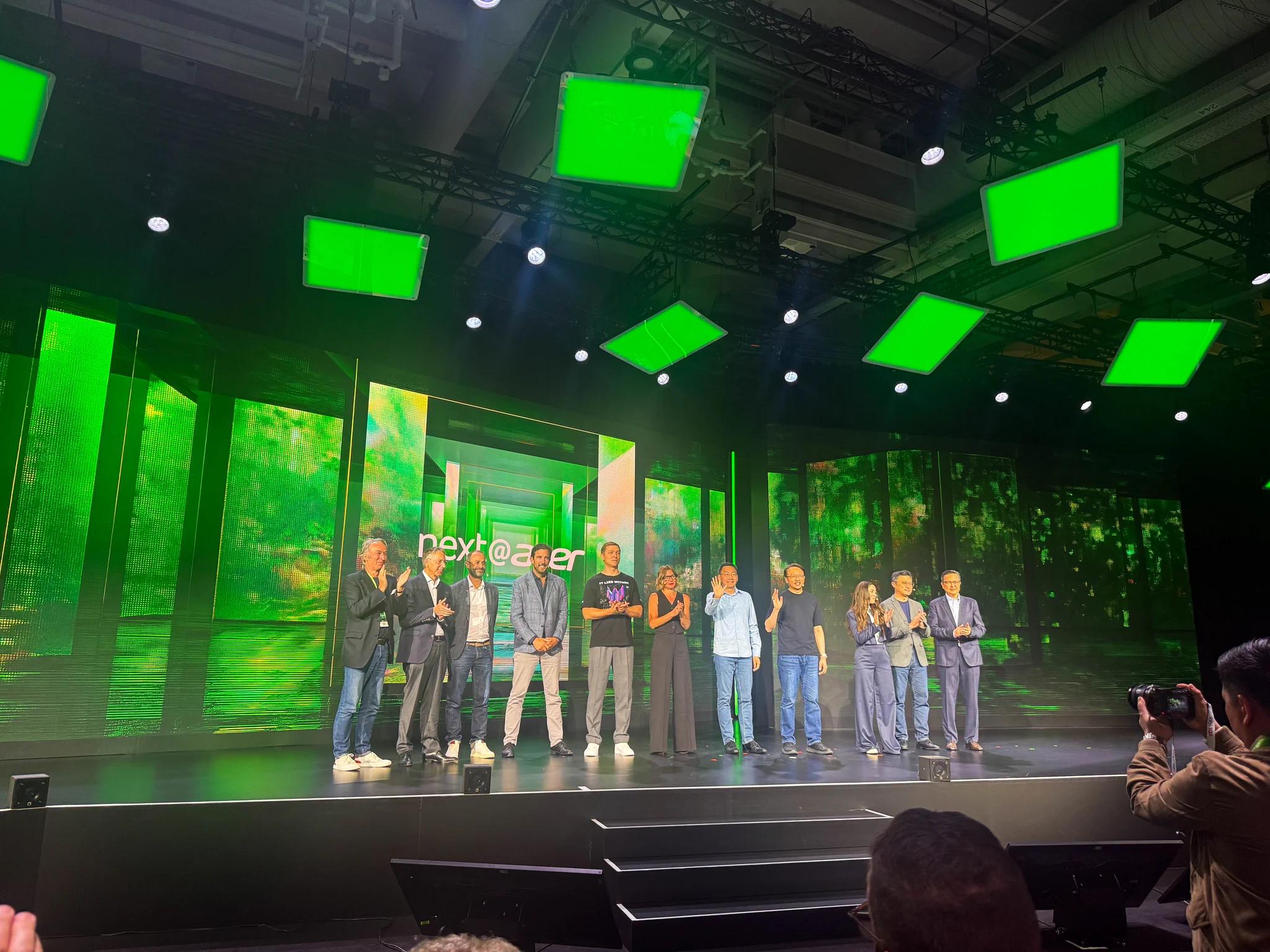 A stage presentation featuring multiple speakers standing together, with a bright green and nature-themed backdrop.