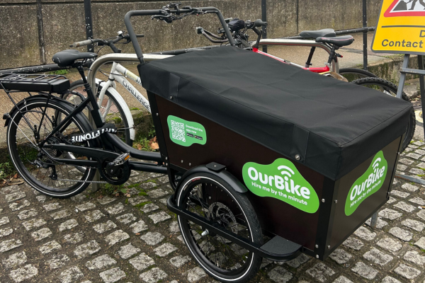 An OurBike cargo e-bike with a black storage box parked on a cobblestone street, with other bicycles in the background.