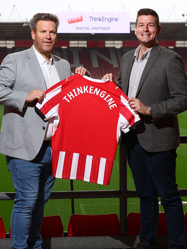 Tom Barrett, Head of Partnerships at Southampton and Ben Michaelis, Managing Director at ThinkEngine, hold up a Saints shirt between them with 'ThinkEngine' printed on the back of the shirt.