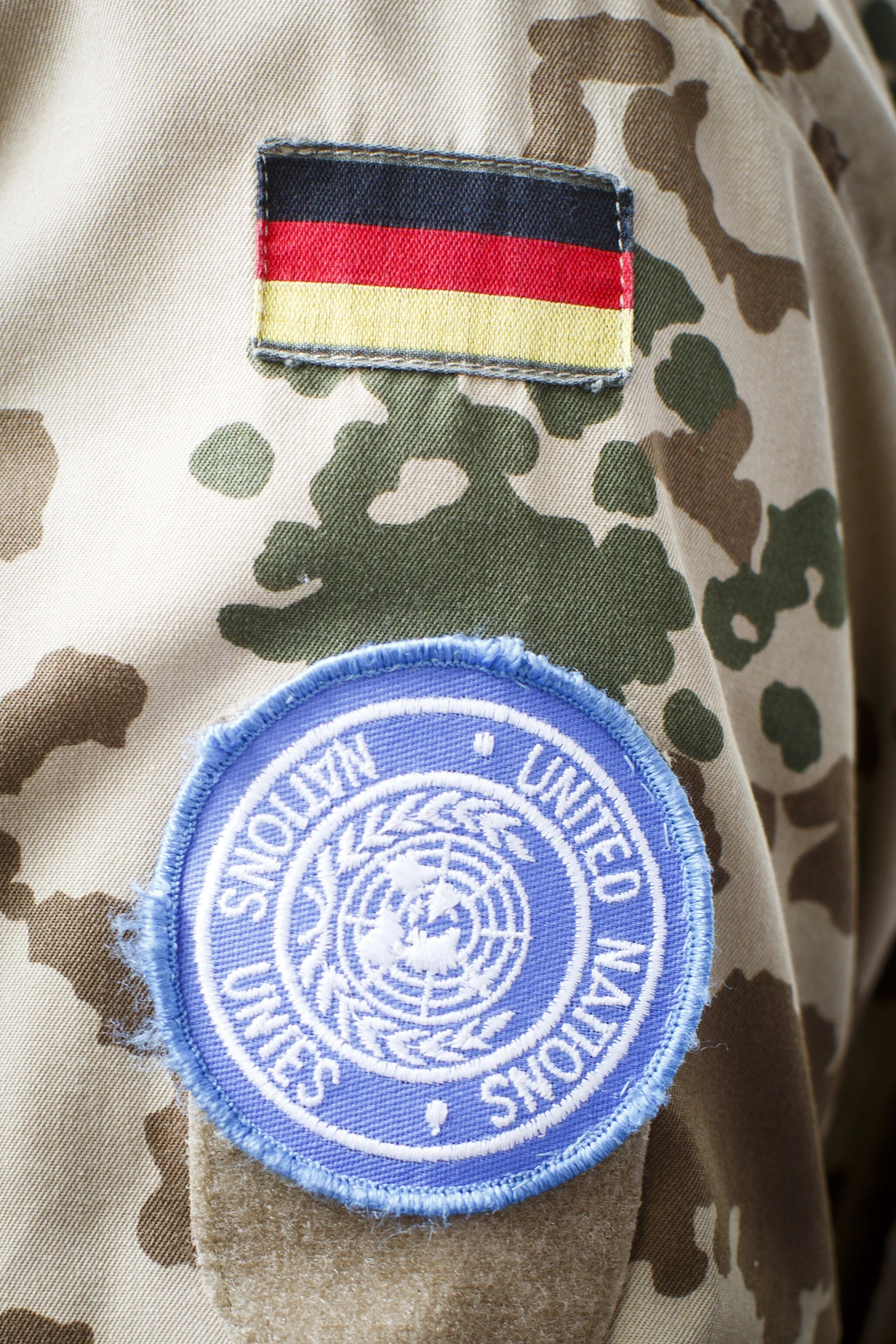 A camouflage-patterned fabric displays two patches: one with horizontal stripes in black, red, and yellow, and another blue circular patch with the words "United Nations" encircling a white emblem.