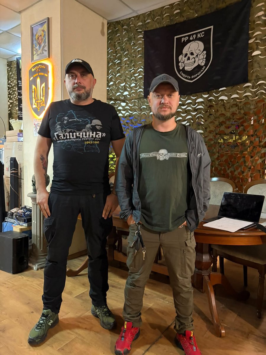 HavryshkoMarta's tweet photo. On the normalization of Nazi symbols in the Ukrainian military.

In the photo, you can see Mykhailo Stoikevych, Chief of Staff of the 49th Assault Battalion “Karpatska Sich,” wearing a T-shirt glorifying the Waffen-SS Galicia Division. 

Next to him is battalion officer Ihor Kostiuk, wearing a shirt bearing the symbol of the 3rd SS Panzer Division “Totenkopf.” And, just in case the dress code was too subtle, the same symbol is hanging on the wall.

Judging by the surroundings, this appears to be some kind of battalion headquarters. Apparently, interior design has taken a rather… historically adventurous turn.

So what does Ukraine’s Jewish Commander-in-Chief, Volodymyr Zelenskyy, have to say about this? And what will The @Jerusalem_Post say? Will it somehow fail to notice it again?