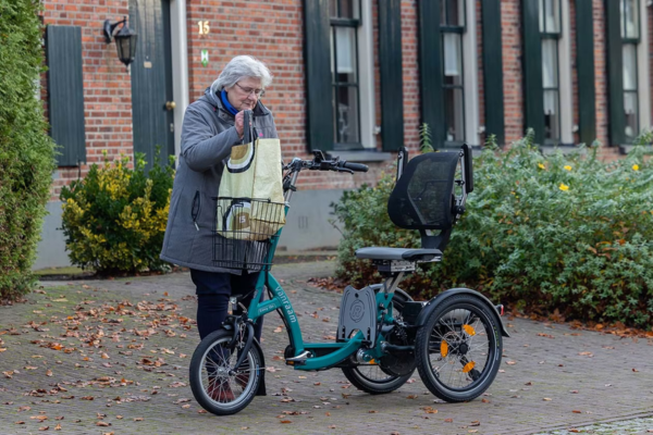 An elderly woman placing a shopping bag into the basket of a teal-coloured Van Raam tricycle on a paved driveway.
