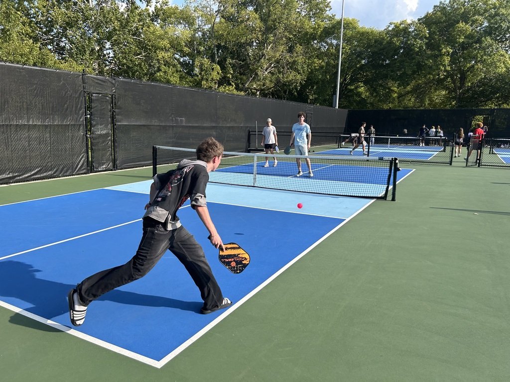 An outdoor tennis court with several players. One player swings a racket to hit a ball.