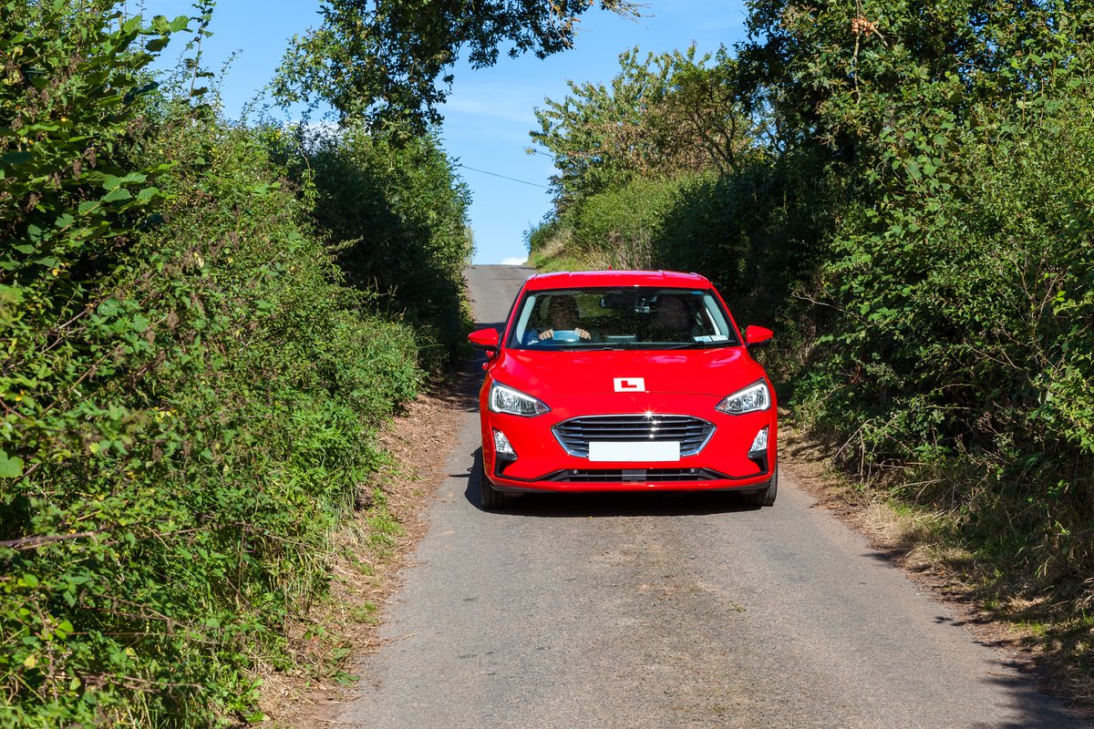 Red learner driver car on a narrow country road surrounded by dense green hedges under a clear blue sky.