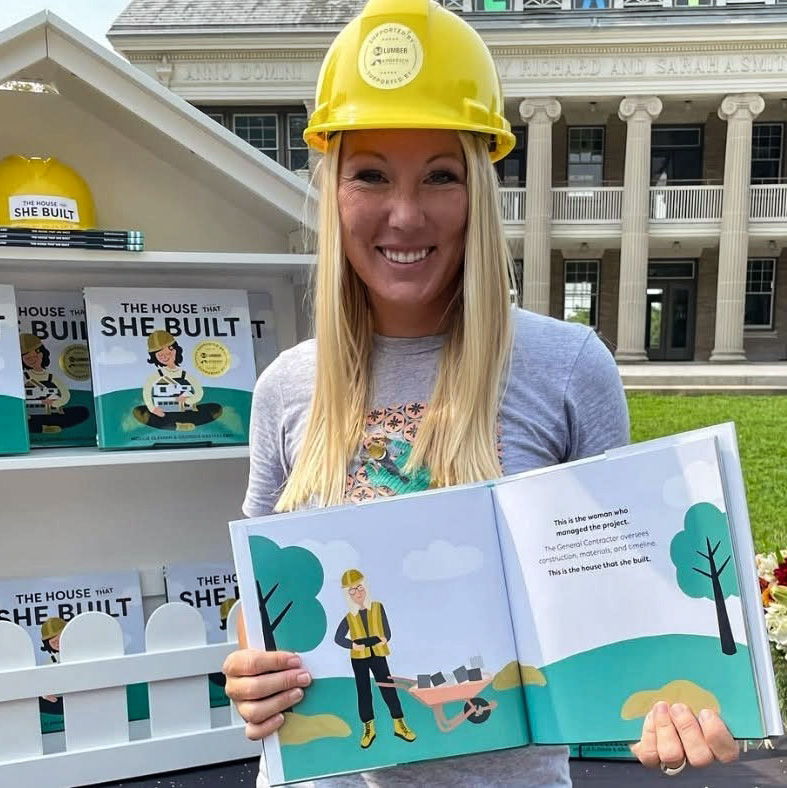 Woman in a hard hat holding an open book with book displays behind her.