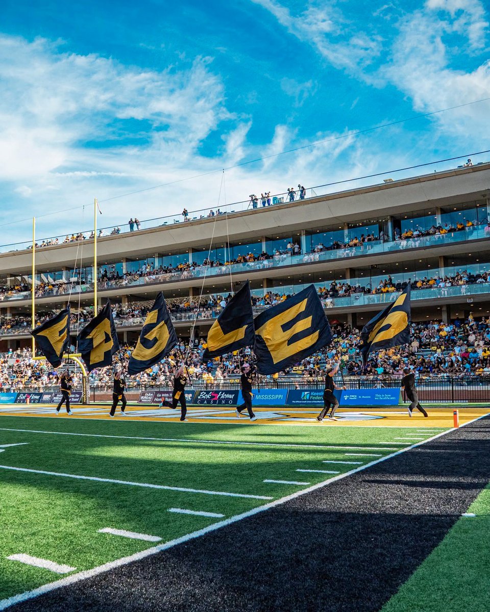 Under a bright blue sky, fans fill the stands during a Southern Miss football game. Several Southern Miss cheerleaders run on the field each holding a black flag with letters spelling out "Eagles."