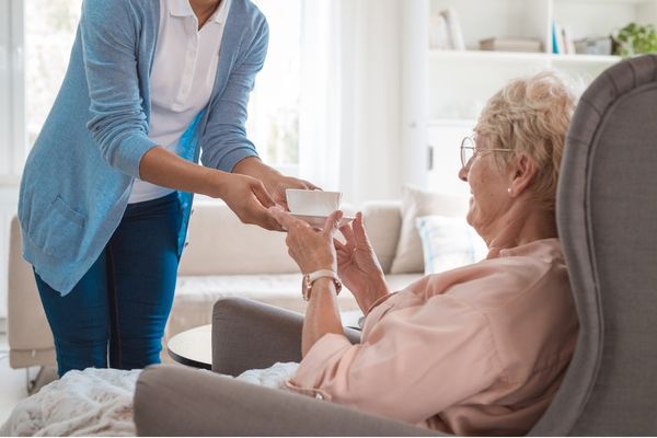 A caregiver in a blue cardigan hands a cup of tea to an elderly woman sitting in an armchair.