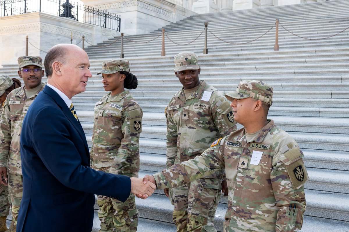 A man in a dark suit shakes hands with a U.S. Army soldier in camouflage while several other soldiers stand nearby on the steps of a large stone building. Uniform patches and name tags are visible.