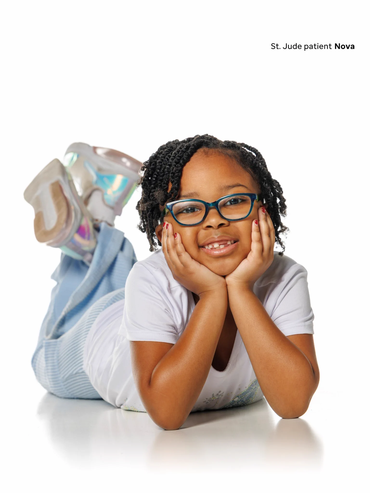 St. Jude patient Nova, a young girl with braided hair and teal glasses, smiles brightly at the camera while lying on her stomach, propping her head with her hands. She wears a white tee, striped blue pants, and iridescent sneakers.