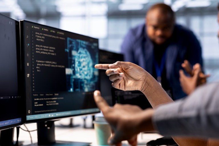 Ambiente de trabalho tecnológico onde duas pessoas interagem em primeiro plano diante de um monitor de computador. A pessoa à direita, em primeiro plano, aponta com o dedo indicador para a tela, enquanto outra mão aparece logo abaixo fazendo um gesto semelhante.

O monitor exibe linhas de código de programação à esquerda e a representação gráfica azul de um cérebro digital à direita. Ao fundo, fora de foco, um homem negro de camisa azul observa a discussão. A iluminação é moderna e o ambiente remete a um escritório de desenvolvimento de software ou inteligência artificial. Imagem de DC Studio no Magnific.