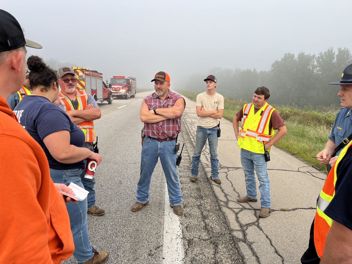 Emergency responders and utility workers gather on a foggy road beside fire trucks, discussing a roadside incident.