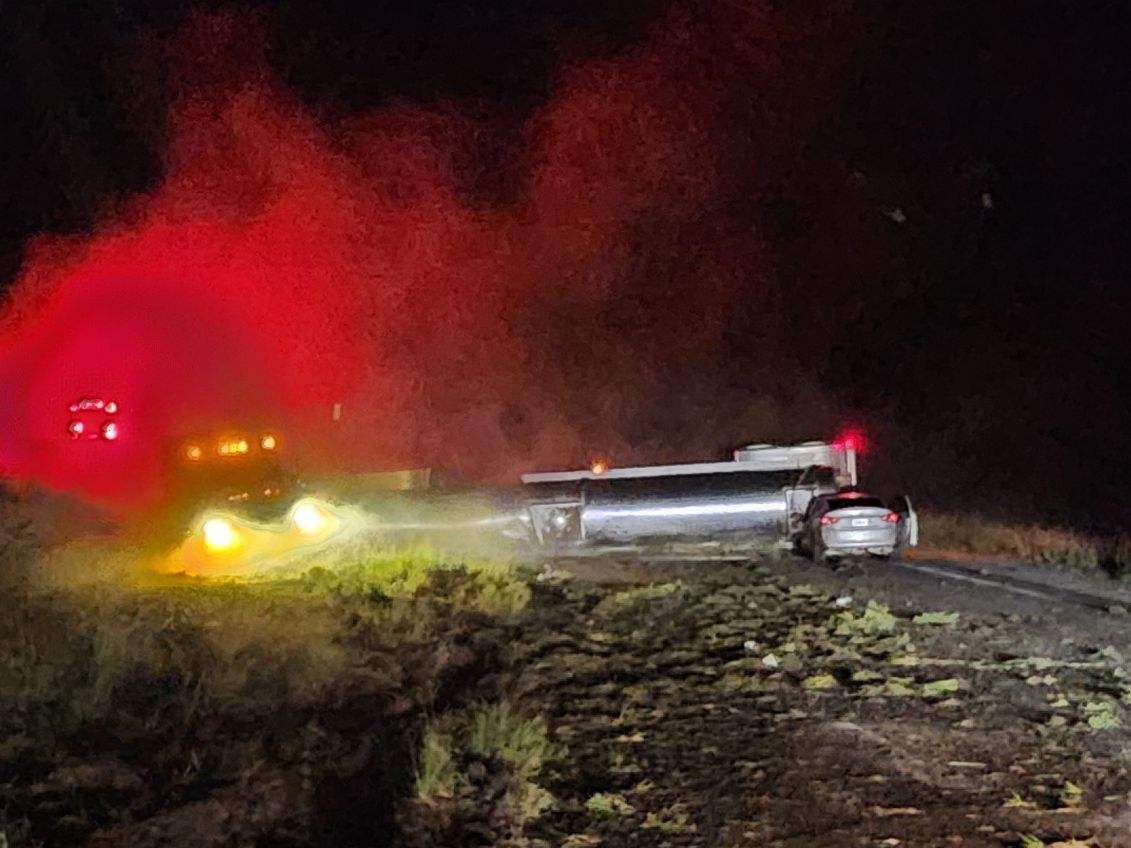 A tanker truck overturned on a dark rural road with emergency vehicle lights illuminating red and yellow smoke in the background.