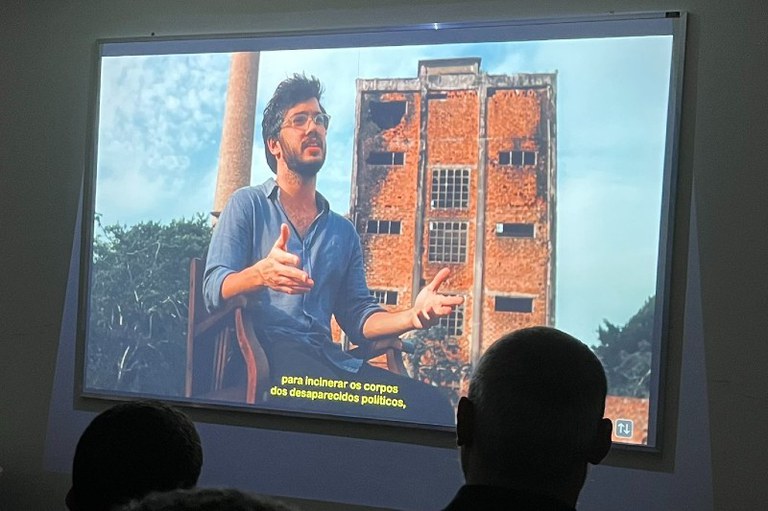 Fotografia de uma projeção numa sala escura. A imagem projetada mostra um homem jovem, de cabelo escuro, barba e óculos, a usar uma camisa azul, sentado numa cadeira de madeira e a gesticular com as mãos abertas enquanto fala. Ao fundo da cena projetada, vê-se a estrutura em tijolo e cimento de um edifício antigo e em ruínas, além de uma chaminé alta do lado esquerdo. Na parte inferior da projeção, surgem legendas em amarelo com o texto: "para incinerar os corpos dos desaparecidos políticos,". Foto: Comunicação/MPF/RJ.